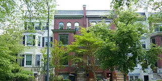 An image of a group of brick rowhouses in Washington, D.C. The Hurd House is the one in the middle, a red brick exterior with a green tree in front partially obscuring the facade. 