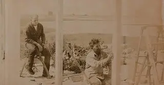 A sepia toned image with a man working on constructing some walls while another man sitting behind him observing with a cigarette in his hand. The background is of a bay of water and an open landscape.. 