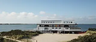 A wide angle view of a beached barge with  three colorful squares on one of the upper stories and a ramp leading up to the entrance. It is white and black with a small beach in front and the expanse of the bay behind it. 
