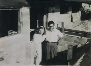 Black and white image of a man and a woman sitting inside a home that is under construction. Mabel is leaning over,  with wide smile across her face, and Victor has a hand on his hip looking forward. 