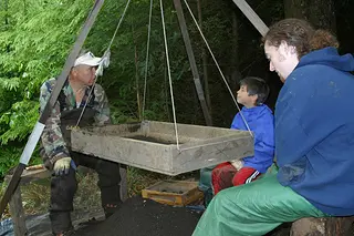 A view of an archaeological field school. The instructor sits on one side of a sieve with two other individuals, students, on the other side. 
