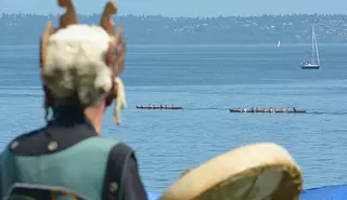 A view of an individual in traditional Suquamish clothing looking out over a body of water where two canoes are racing. There is a sailboat in the background. 
