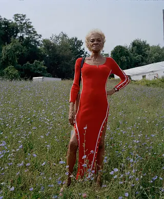 A woman standing in a field of purple flowers wearing a bright red dress with white piping along the sleeves and the edges. 